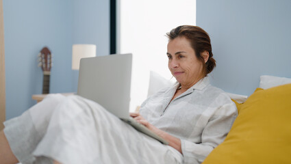 Middle age hispanic woman using laptop sitting on bed at bedroom
