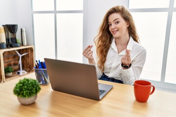 Young caucasian woman working at the office using computer laptop doing money gesture with hands,...