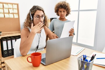 Young business mother working at the office with kid covering mouth with hand, shocked and afraid...