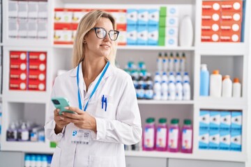 Young blonde woman pharmacist using smartphone working at pharmacy