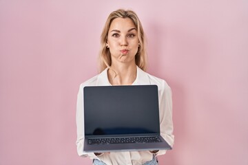 Young caucasian woman holding laptop showing screen puffing cheeks with funny face. mouth inflated...