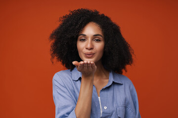 Young middle-eastern woman with curly hair blowing air kiss at camera - obrazy, fototapety, plakaty