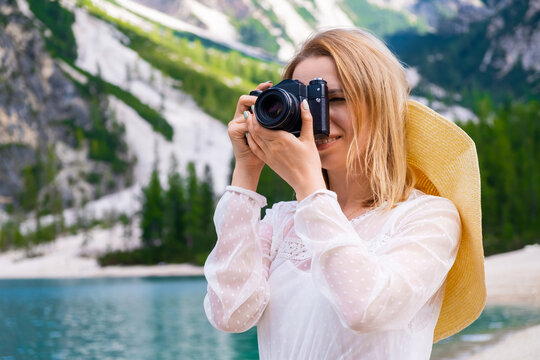 Smiling blonde hair female photographer with a professional camera taking photo of the lake. Portrait of woman in summer hat and white dress against Dolomites alps. 