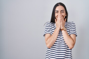 Young brunette woman wearing striped t shirt laughing and embarrassed giggle covering mouth with...