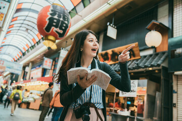 excited asian female visitor spotting landmark and finger pointing at distance while using guidebook in kuromon ichiba market in Osaka japan. translation on lantern:â kuromonâ