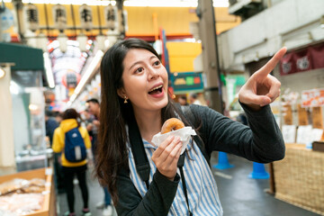 excited asian female traveler noticing something fun and finger pointing at distance while having baked doughnut for snack in kuromon ichiba market in Osaka japan