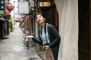 Fototapeta na wymiar asian female traveler with camera looking up at sky and reaching out hand to see if itâs raining outside of Japanese restaurant in a tradition alleyway near nanba-eki Osaka japan