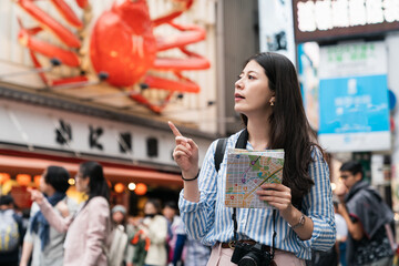lost Chinese girl sight-seer finger pointing at space while consulting a map to find out where she is in shinsaibashi suji and doutonbori shopping district in Osaka japan