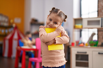 Adorable hispanic girl student hugging book standing at kindergarten