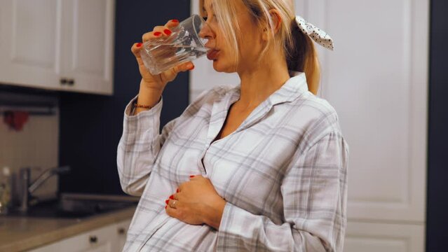 Happy peaceful attractive young pregnant woman drinking glass of fresh pure mineral water, preventing dehydration enjoying daily healthcare habit, stroking big belly, healthy pregnancy concept. 