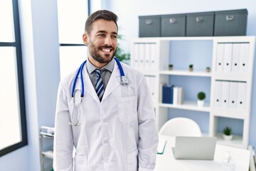 Handsome hispanic man wearing doctor uniform and stethoscope at medical clinic looking away to side with smile on face, natural expression. laughing confident.