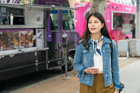 Asian Woman Holding Smartphone And Looking Away Is Seeking Food For Lunch. Korean Lady Just Moving And Living Alone Overseas Is Trying To Get Familiar With The Environment.