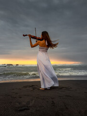 Caucasian woman with violin on the beach. Music and art concept. Slim girl wearing long white dress and playing violin in nature. Sunset time. Cloudy sky. View from back. Bali © Olga
