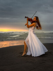 Caucasian woman with violin on the beach. Music and art concept. Slim girl wearing long white dress and playing violin in nature. Sunset time. Cloudy sky. Bali