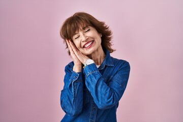 Middle age woman standing over pink background sleeping tired dreaming and posing with hands together while smiling with closed eyes.