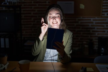 Blonde caucasian woman working at the office at night gesturing finger crossed smiling with hope and eyes closed. luck and superstitious concept.