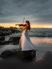 Charming Caucasian woman with violin on the beach. Music and art concept. Slim girl wearing long white dress and playing violin in nature. Sunset time. Cloudy sky. Bali