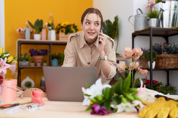 Young caucasian woman florist talking on smartphone using laptop at flower shop