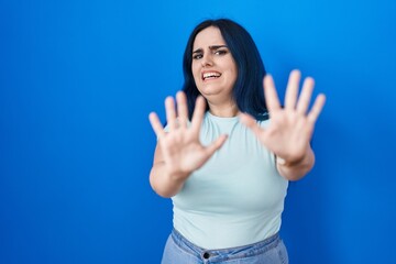 Fototapeta premium Young modern girl with blue hair standing over blue background afraid and terrified with fear expression stop gesture with hands, shouting in shock. panic concept.