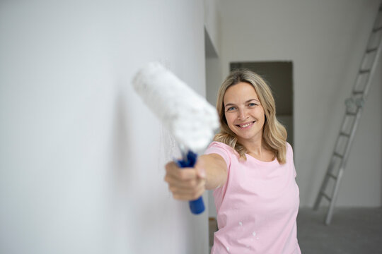 Smiling woman holding paint bucket standing near white wall in apartment