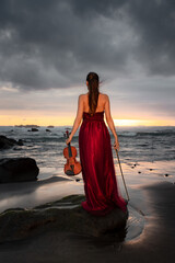 Caucasian woman with violin on the beach. Music and art concept. Slim girl wearing long red dress and holding violin in nature. Sunset time. Cloudy sky. View from back. Bali