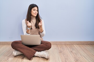 Young brunette woman working using computer laptop sitting on the floor doing money gesture with hands, asking for salary payment, millionaire business