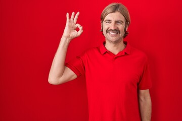 Caucasian man with mustache standing over red background smiling positive doing ok sign with hand and fingers. successful expression.