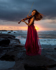Charming Caucasian woman with violin on the beach. Music and art concept. Slim girl wearing long red dress and playing violin in nature. Sunset time. Cloudy sky. Bali © Olga