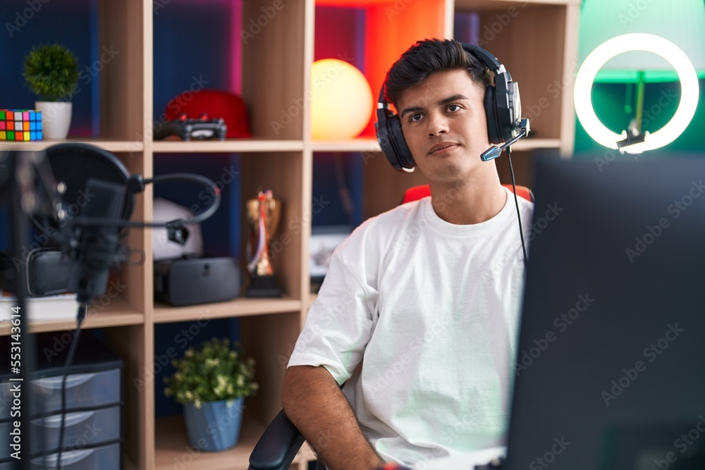 Sticker Young hispanic man streamer sitting on table with relaxed expression at gaming room