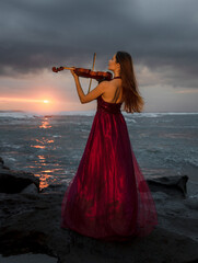 Charming Caucasian woman with violin on the beach. Music and art concept. Slim girl wearing long red dress and playing violin in nature. Sunset time. Cloudy sky. Bali