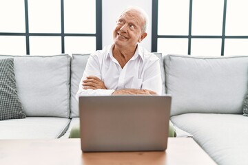 Senior man using laptop at home sitting on the sofa smiling looking to the side and staring away thinking.