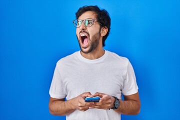 Handsome latin man using smartphone typing message angry and mad screaming frustrated and furious, shouting with anger. rage and aggressive concept.