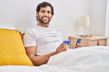 Obraz premium Young hispanic man using smartphone and credit card sitting on bed at bedroom