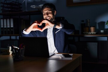 Handsome latin man working at the office at night smiling in love showing heart symbol and shape with hands. romantic concept.