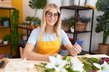 Young blonde woman florist using smartphone holding credit card at flower shop