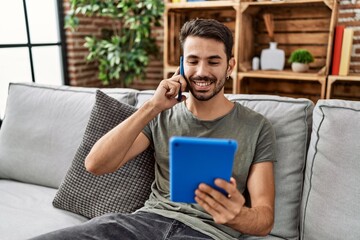 Young hispanic man using touchpad and talking on the smartphone at home