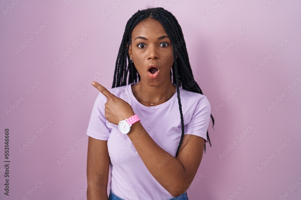 Canvas Prints African american woman with braids standing over pink background surprised pointing with finger to the side, open mouth amazed expression.