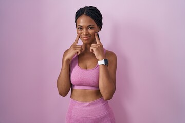 Obraz na płótnie Canvas African american woman with braids wearing sportswear over pink background smiling with open mouth, fingers pointing and forcing cheerful smile