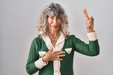 Middle age woman standing over white background smiling swearing with hand on chest and fingers up,...
