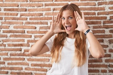Young caucasian woman standing over bricks wall smiling cheerful playing peek a boo with hands showing face. surprised and exited