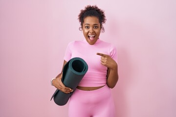 Young hispanic woman with curly hair holding yoga mat over pink background surprised pointing with finger to the side, open mouth amazed expression.
