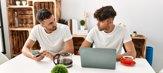 Two hispanic men couple using smartphone and laptop at home