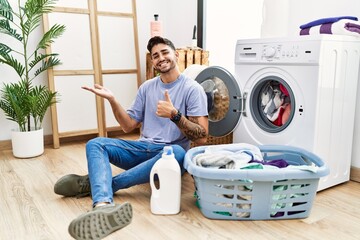 Young hispanic man putting dirty laundry into washing machine showing palm hand and doing ok gesture with thumbs up, smiling happy and cheerful