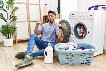 Young hispanic man putting dirty laundry into washing machine pointing down with fingers showing advertisement, surprised face and open mouth