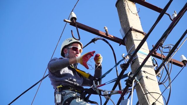 Skilled electrician in helmet fixes wires standing on ladder near high pole against blue sky on summer day backside view. Electrical service and mounting on the pole. Slow motion