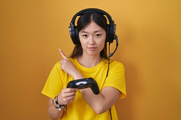 Chinese young woman playing video game holding controller pointing with hand finger to the side showing advertisement, serious and calm face