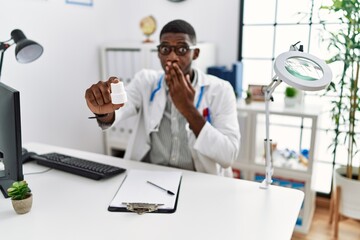 Young african american doctor man wearing doctor uniform holding prescription pills at the clinic covering mouth with hand, shocked and afraid for mistake. surprised expression