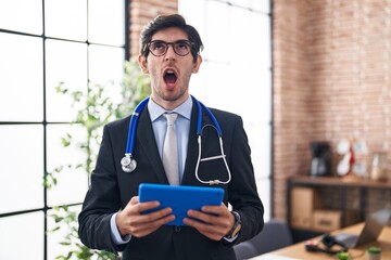 Young hispanic man wearing doctor stethoscope at the office angry and mad screaming frustrated and furious, shouting with anger looking up.