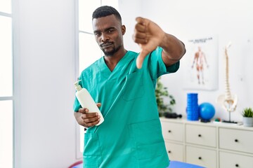 Young african physiotherapist man holding massage body lotion with angry face, negative sign...