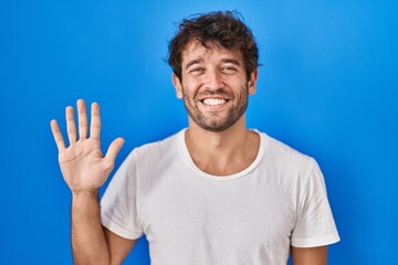Hispanic young man standing over blue background showing and pointing up with fingers number five...